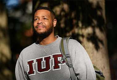 a young man in an IUP sweatshirt stands facing off camera in the oak grove with the shadows of tree branches covering him