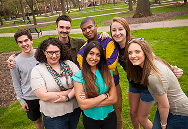a group of young men and women smiling for the camera while grouped together.  In the background is sidewalks and oak trees