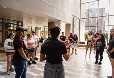 a group of young men and women standing in a lobby as the person in the middle faces away from the camera and speaks to the group