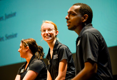 three young students sitting in front of a projection screen while wearing black IUP polo shirts