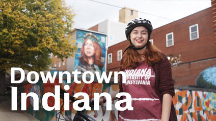 a young woman with a bicycle and wearing a helmet smiles for the camera while backdropped by hand painted murals on building sides