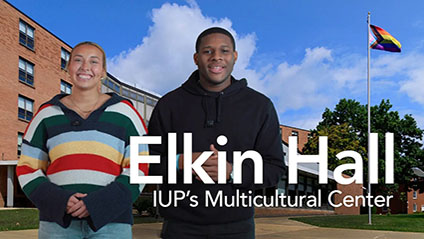 A female and male student stand outside of a brick building with a progress pride flag flying overhead.