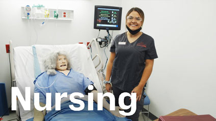 a young woman in nursing scrubs smiles for the camera while standing next to a hospital bed containing a training dummy