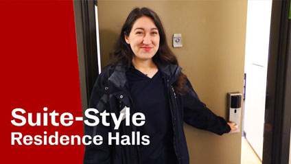 a young woman smiling for the camera while opening a door to a dorm building