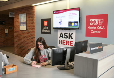 a young woman sits behind a desk writing on a piece of paper in front of signs that say Ask Here and IUP Hawk Q and A center