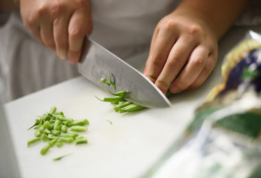 a close up shot of a white cutting board with two hands using a chef's knife to chop the ends off green beans