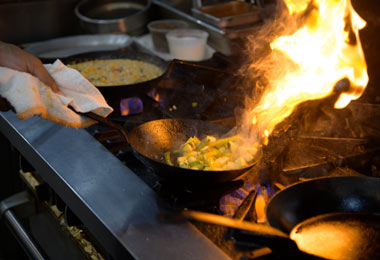 A frying pan with a cloth wrapped around the handle.  A hand is flipping the flaming food within the pan over a hot range.