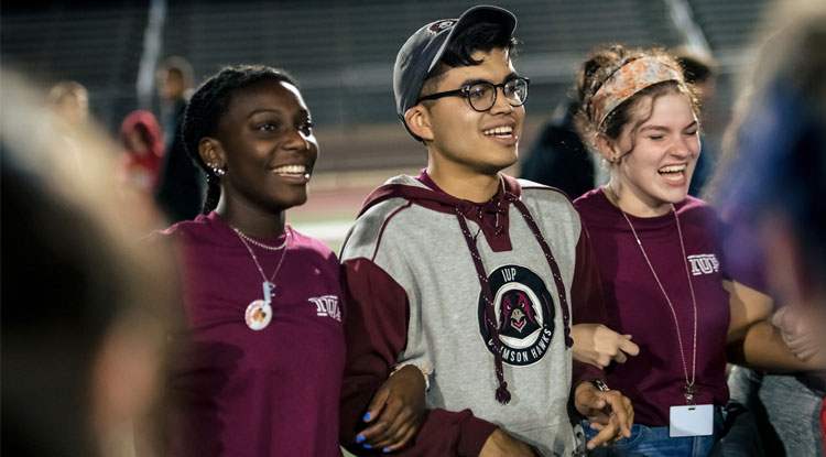 Three students wearing crimson colors stand side by side with their arms interlocked facing to the right