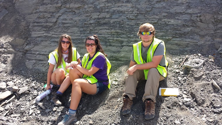 Three students taking a break at the base of a rock outcrop during a field trip.