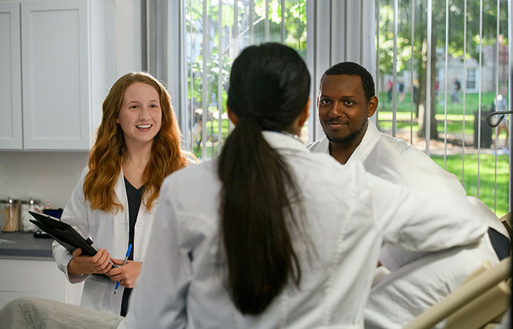 Three students wearing white lab coats talk with each other in a room next to a window