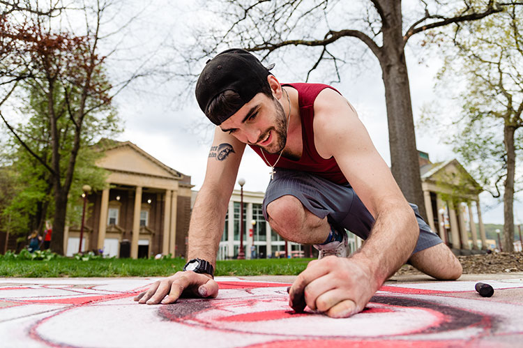 A student chalking the walk in the Oak Grove with the IUP Performing Arts Center in the background