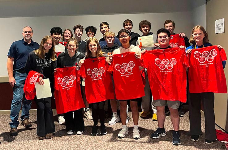 Group photo of Physics Olympics students holding up their event t-shirts, red with the Physics Olympics logo
