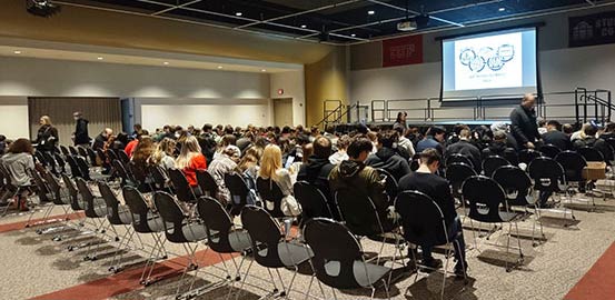 Students sitting in an auditorium with the Physics Olympics logo projected on a screen at the front