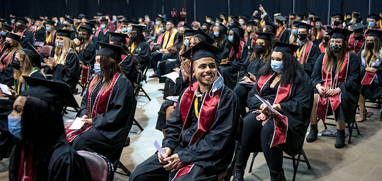 A graduating student smiles while sitting in a chair with other graduates on the floor of the KCAC