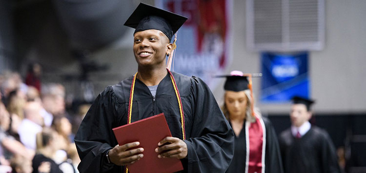 A graduating student carrying their IUP diploma smiles and looks at the audience in the KCAC.