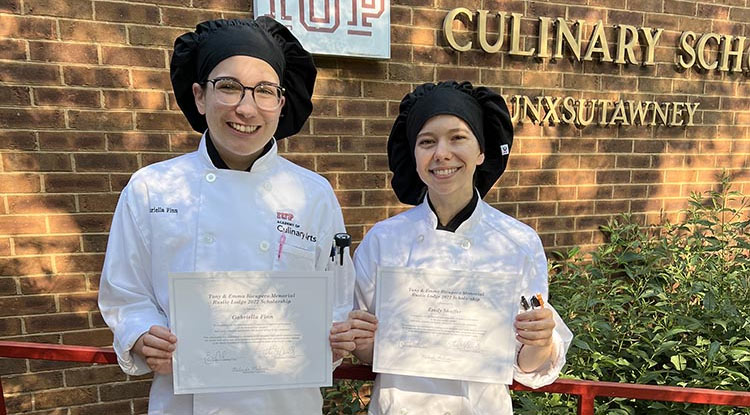 two students in chef outfits holding certificates standing side by side in front of a brick wall