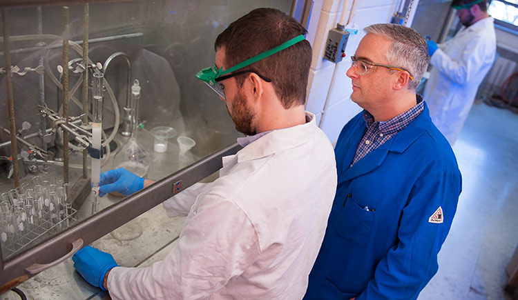 student and professor wearing protective gear, conducting a chemical experiment in the lab standing behind the safety glass divider.