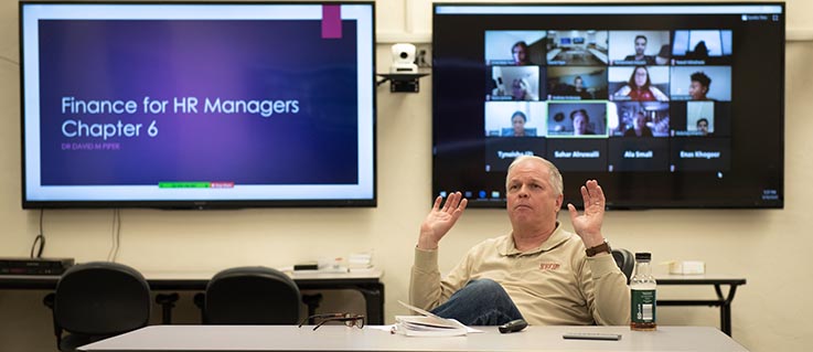  Dr. Piper teaches to a live class with other classmates joining via zoom on the screen behind him.