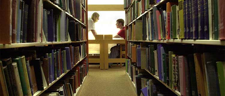 Two IUP students talk at a desk among the library book shelves
