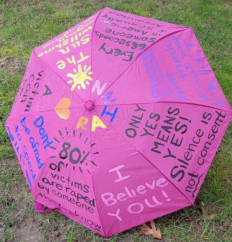 RAINN Day, a open pink umbrella with designs and statistics written on it, in the Oak Grove grass