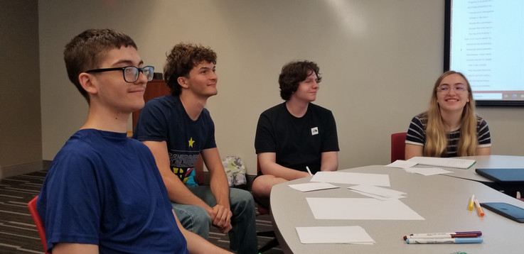 Four students sit at a table. Three are looking at something to the right off camera and one faces the camera.