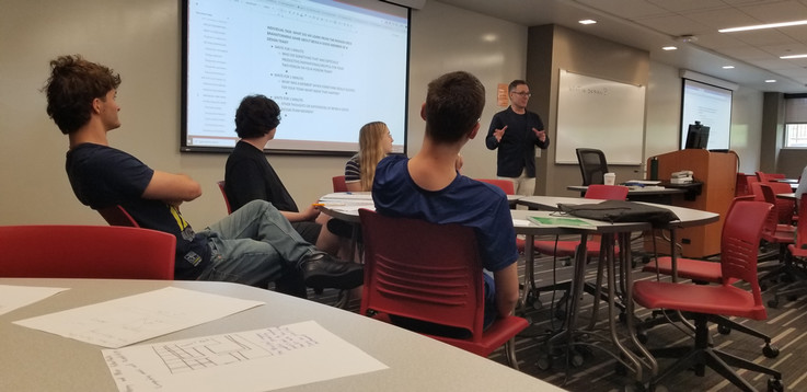 Four students sit at a table. They are turned away from the camera to pay attention and listen as Prof. Sell provides instruction to the class.
