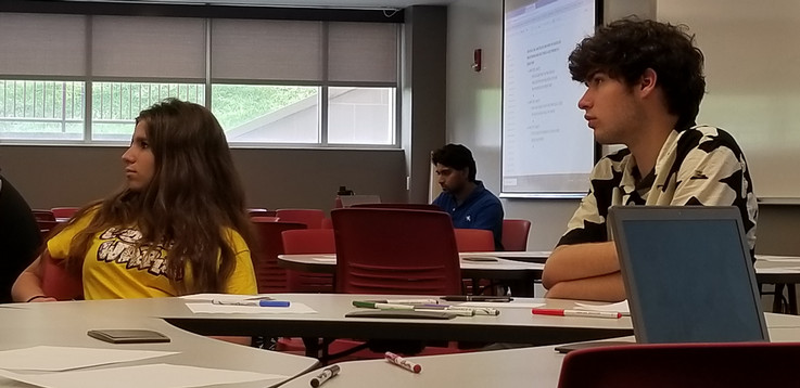 Two students sit a table facing left to look at Prof. Sell off camera and listen as provides instruction. Prof. Sell's grad student is seen in the background between the two students.
