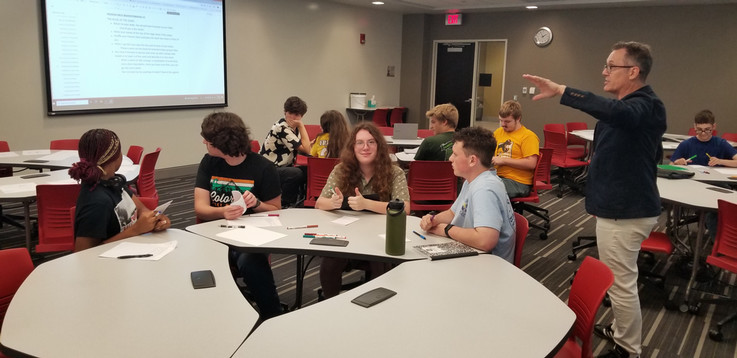 Students sitting at several tables listen as Prof. Sell provides instruction.