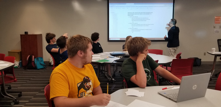 Two students sit at a table. They are turned away from the camera to pay attention and listen as Prof. Sell provides instruction to the class.