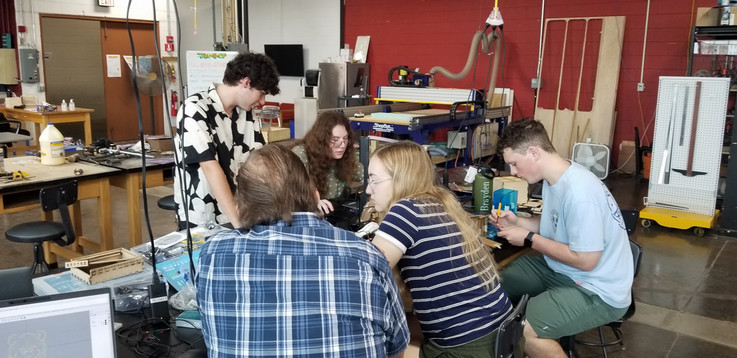 Four students are sitting at work tables in the STEAMSHOP working on their projects. One student is getting guidance from Prof. Adkins.