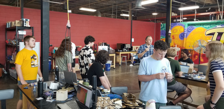 Several students are sitting at and standing around work tables in the STEAMSHOP working on their projects. They are getting guidance from Prof. Adkins.