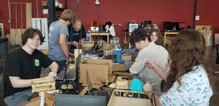Several students are sitting at work tables in the STEAMSHOP working on their projects. One is standing with Prof. Adkins getting guidance on their project. Three students are facing the camera. Two are smiling for the photo.