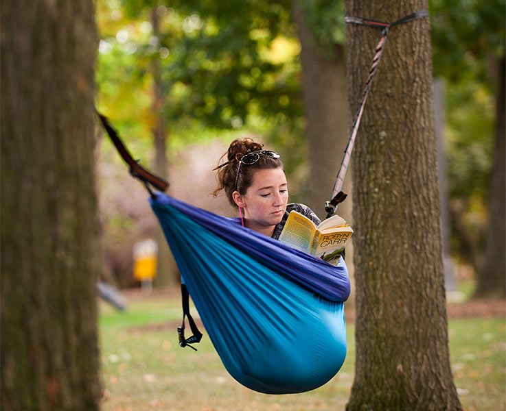 A student reads a book in a hammock hung between trees in the Oak Grove