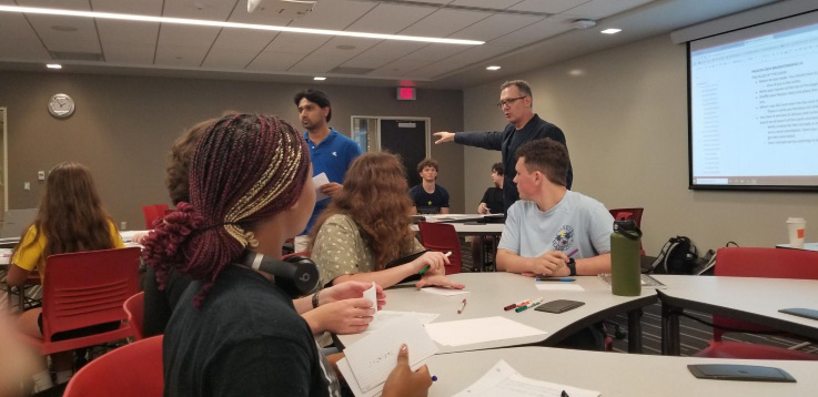 A dozen students sitting at tables in a classroom listen to a professor talk about game design.