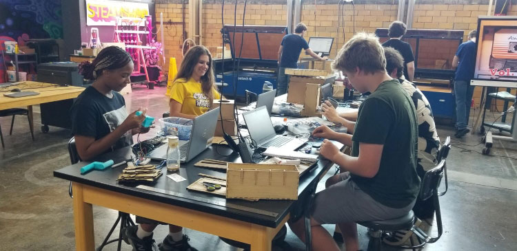 Four high school students in the Cook Honors College Summer Honors Program work on projects at a table covered with tools and laptops in the warehouse-like space housing the IUP STEAMSHOP. In the background, other students use large machines along the back wall to create their projects.