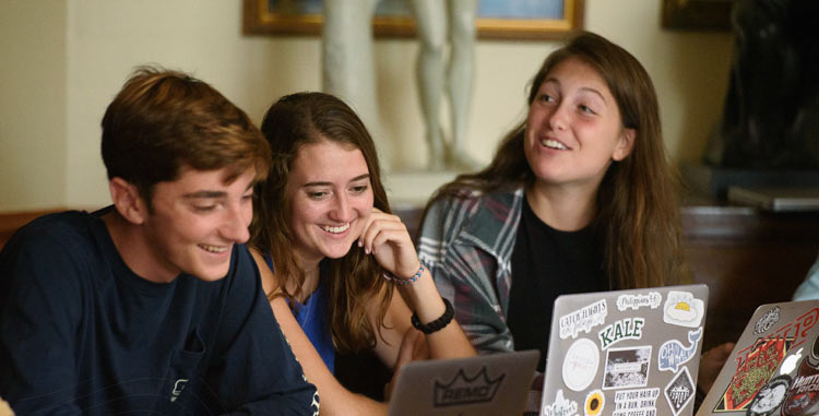 three students sit smiling looking at laptops in the Cook Honors College study room