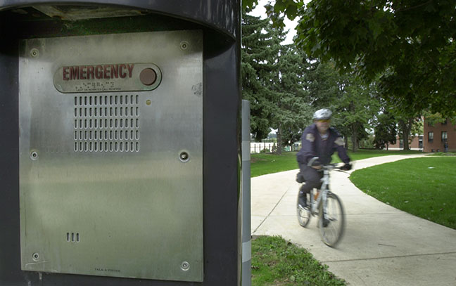 A police officer on a bicycle passes a blue-light phone pillar with an emergency label on it.