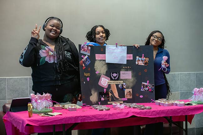 Three students posing at a Valentine's Day-themed table with various wrapped candy for sale