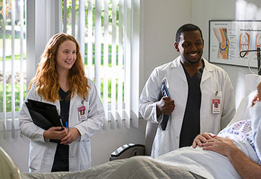 two students in lab coats smiling while standing next to a hospital bed