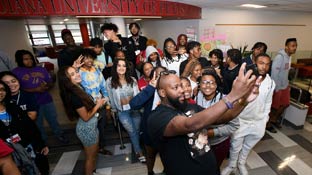 A photo of a young man standing in front of a large group of people.  He has his phone stretched out to take a selfie with everyone.