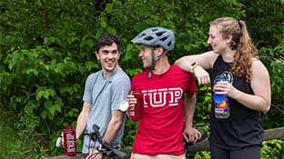 Three students with water bottles stand on a trail with a bicycle. 