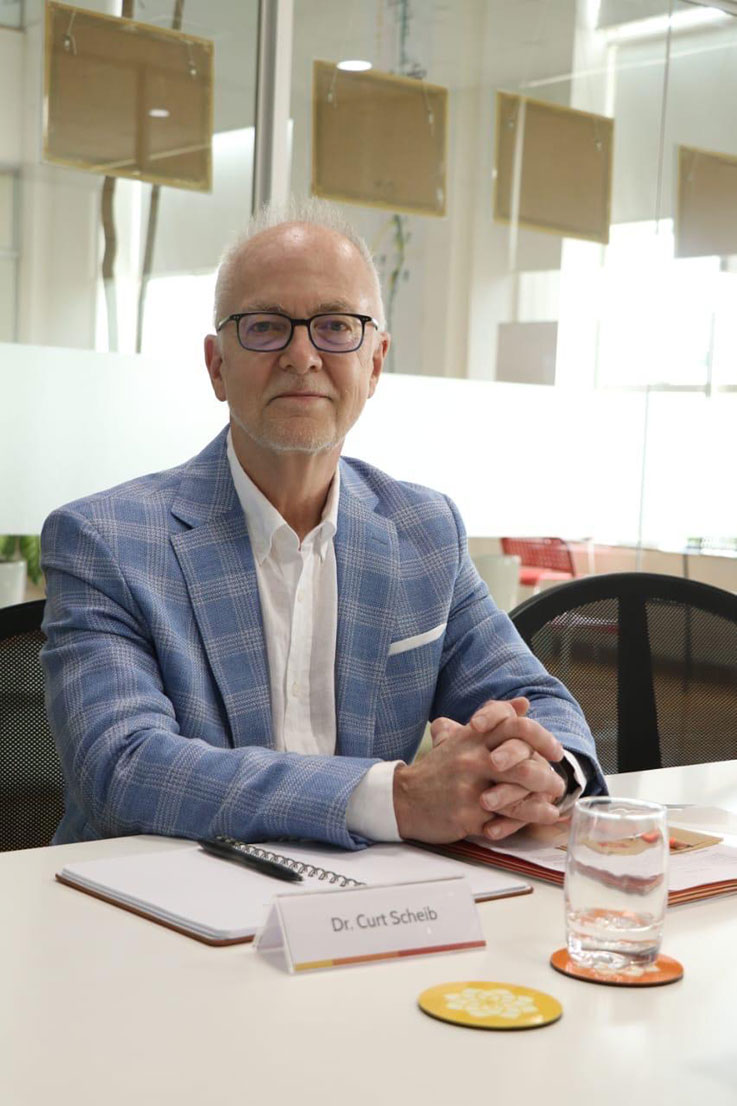 Curt Scheib sits at a table where there is a glass of water, a name card for him and a notebook and pen. He is wearing a white shirt and light blue suit jacket and has glasses on as he smiles for the picture.