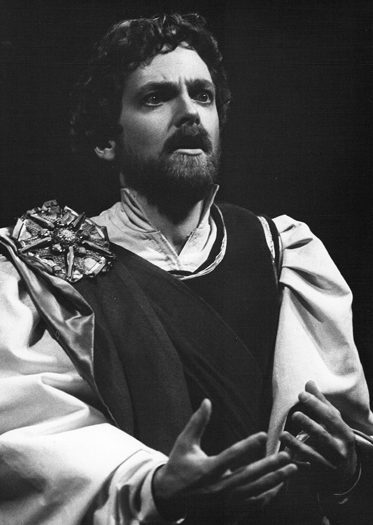 An actor dressed in an elaborate opera shirt with puffed sleeves is photographed in black and white. He has an anxious expression as if the song or lines he is about to deliver are causing him great stress.