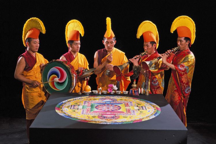 Monks playing Tibetan musical instruments around a completed sand manadala