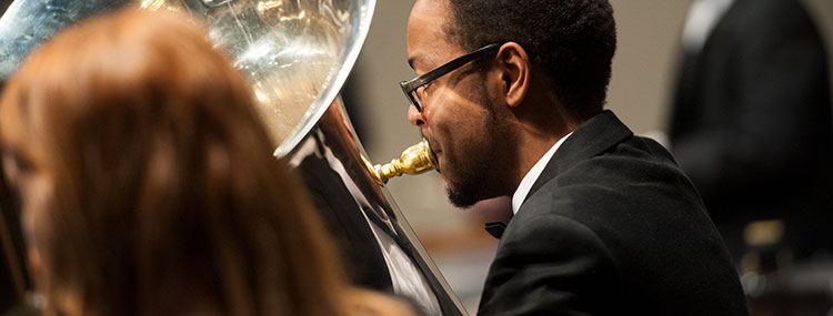 A student dressed in formal black and white clothing plays a tuba in a concert band performance on stage