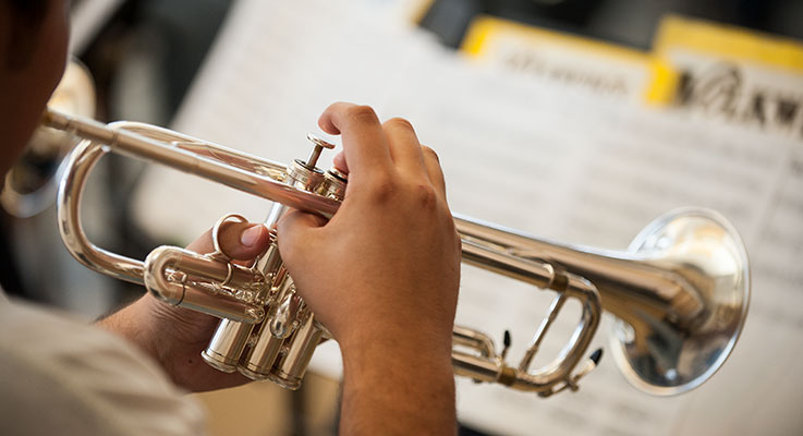  A close-up view of hands playing a trumpet with a music stand holding sheets of music in the background