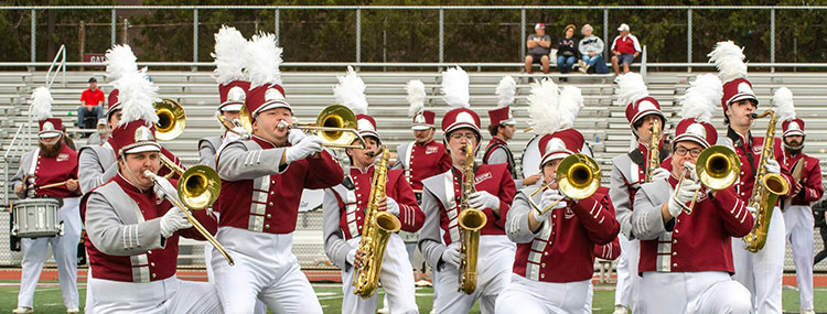 About 15 members of the marching band, dressed in uniforms with Crimson jackets, white pants, and helmets with plumes, perform on a football field