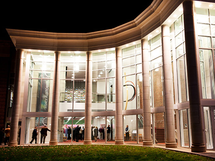 The IUP Performing Arts Center lobby at night with light shining through the tall wall of glass windows.