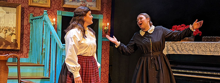Two women in old-fashioned long skirts and cotton shirts perform on stage, one with her arms outstretched, during the musical Little Women