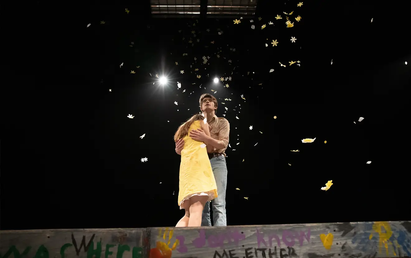A man and woman embracing on a dark stage as leaves fall around them.